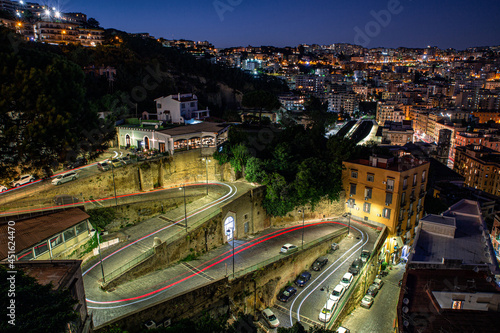 Night view over the streets and lights of the city of Naples with a serpentine road in the foreground with a car pulling light stripes.