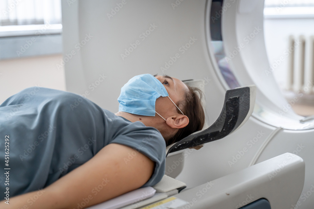Obraz premium A woman in a medical mask lies on the tomograph table. woman is undergoing computed axial tomography examination in a modern hospital.