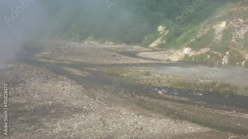 In the Valley of Geysers, a winding river flows along a rocky bed.  There are hot springs nearby, steam flies over the ground. There is green vegetation on the mountain slopes. Kamchatka