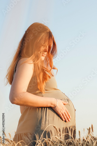 beautiful young pregnant woman walks on wheat field at sunset, expectant mother with relax in nature petting her belly with hand, happy pregnancy concept