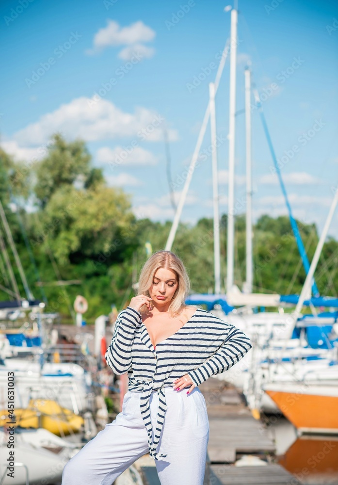 Pretty blonde woman of plus size, American or European appearance near yachts, enjoying life. Young lady with excess weight, stylishly dressed at striped jumper and white pants .Natural beauty Pretty blonde woman of plus size, American or European appearance near yachts, enjoying life. Young lady with excess weight, stylishly dressed at striped jumper and white pants .Natural beauty