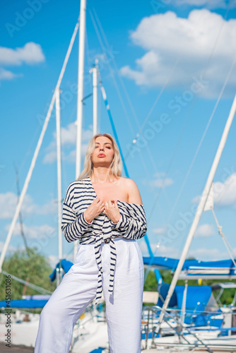 Pretty blonde woman of plus size, American or European appearance near yachts, enjoying life. Young lady with excess weight, stylishly dressed at striped jumper and white pants .Natural beauty