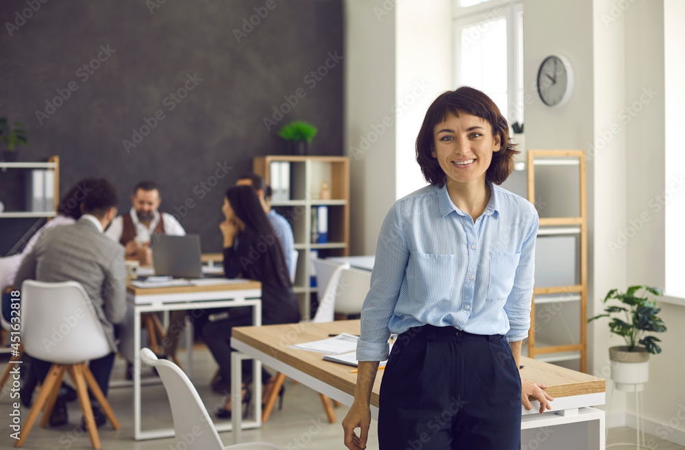Happy beautiful young European woman standing in a room of a modern office workspace. Portrait of a successful female business executive or company worker leaning on a table and looking at the camera Happy beautiful young European woman standing in a room of a modern office workspace. Portrait of a successful female business executive or company worker leaning on a table and looking at the camera