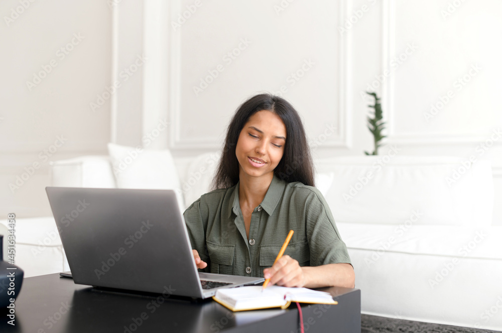 Freelance woman using laptop for remote work from home. Indian female student in casual wear sits at the desk, watching online webinars or training and taking notes, writes down Freelance woman using laptop for remote work from home. Indian female student in casual wear sits at the desk, watching online webinars or training and taking notes, writes down