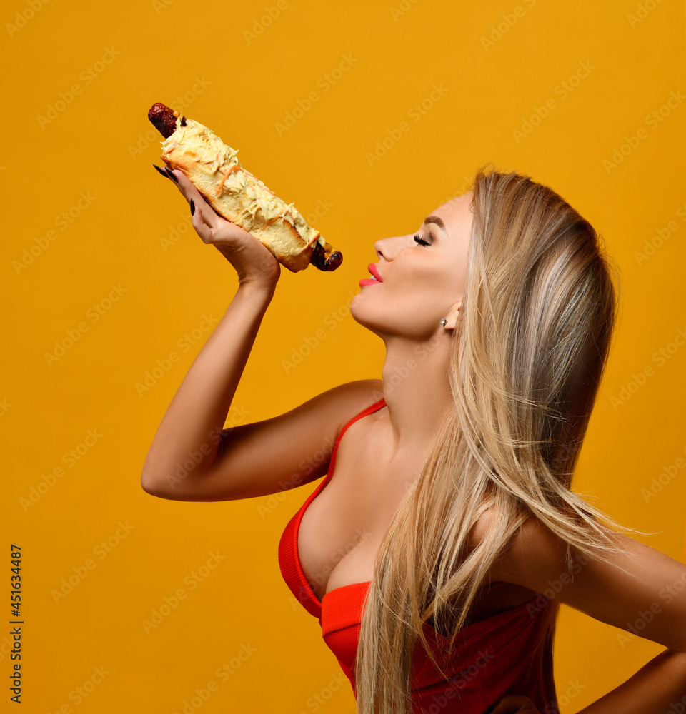 Sexy blonde woman holds a delicious hot dog, smiling happily surprised,  laughing in a red bikini on a yellow background. Fast or junk food concept.  Stock-Foto | Adobe Stock