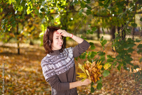 Happy caucasian young woman in a sweater holds bright autumn leaves in her hand. Happy smiling girl in the park. Seasonal autumn concept