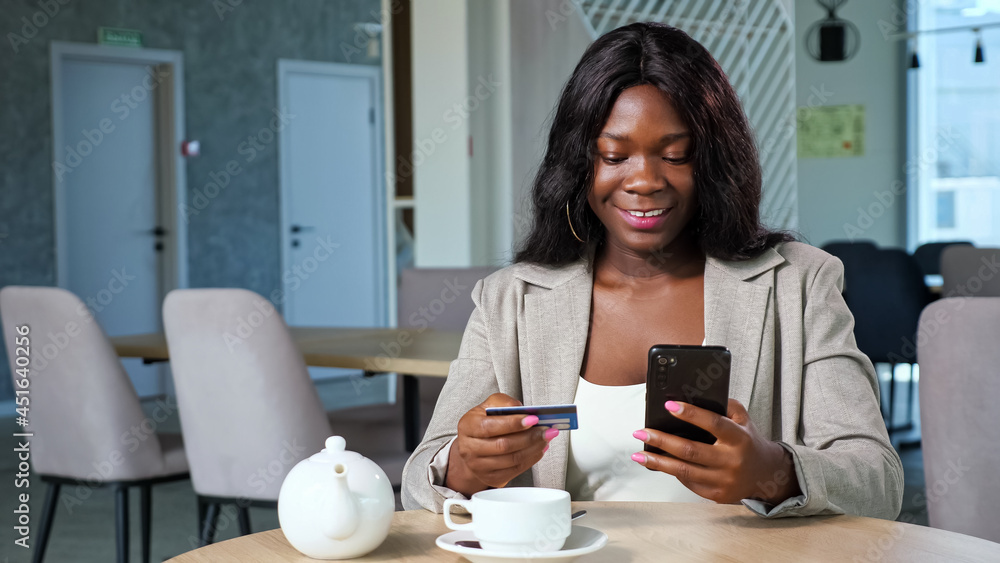 Cheerful black woman in stylish jacket pays online with credit card and mobile phone sitting at table with teapot and cup in cozy cafeteria Cheerful black woman in stylish jacket pays online with credit card and mobile phone sitting at table with teapot and cup in cozy cafeteria