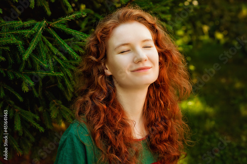 A large portrait of a beautiful woman with long red hair against the background of greenery in the park. Her eyes are peacefully closed  the woman enjoys nature.