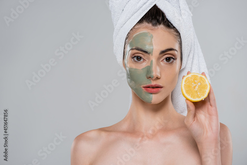 young woman with towel on head and green clay mask on face holding half of orange isolated on grey