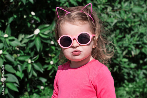 a little girl with blonde hair wearing pink glasses and a pink dress. funny little baby in pink in summer on the street