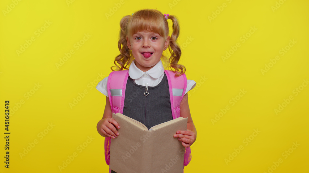 Smiling schoolgirl wears backpack, holding a book, laugh fooling around showing tongue making playful silly facial expressions and grimacing over yellow studio background. Back to school concept Smiling schoolgirl wears backpack, holding a book, laugh fooling around showing tongue making playful silly facial expressions and grimacing over yellow studio background. Back to school concept