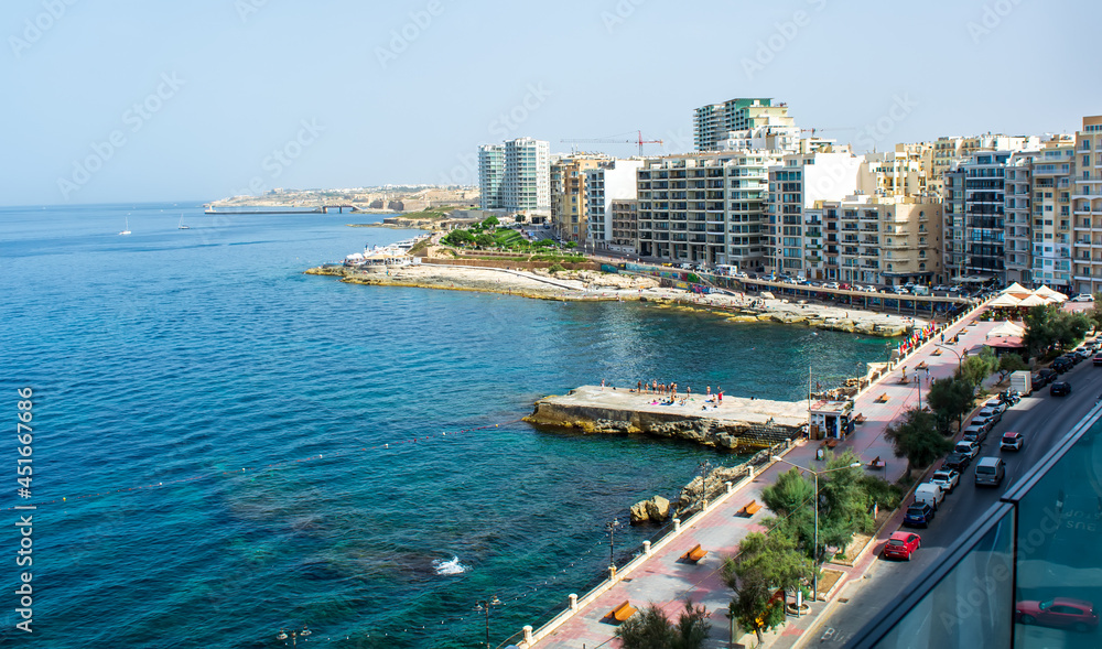Sliema rocky coastline, Malta, with cafes, modern buildings, and bridge at the distance.