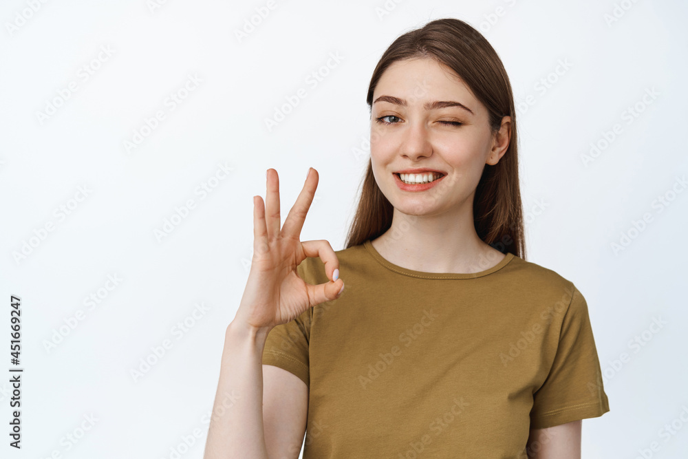 Winking girl shows okay, OK sign and smiling, nod in approval, recommending something good, compliment quality, white background Winking girl shows okay, OK sign and smiling, nod in approval, recommending something good, compliment quality, white background