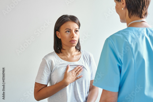 Worried young afro american woman patient touching chest, explaining symptoms and complaining about heartache to female doctor in blue uniform while visiting clinic office. Professional medical care