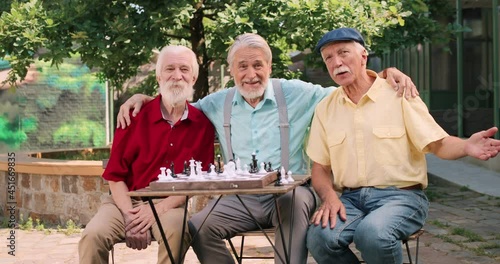 Waist up portrait view of the old caucasian man spending time in his yard with his two old best friend while playing chess and embracing. Outdoor. photo