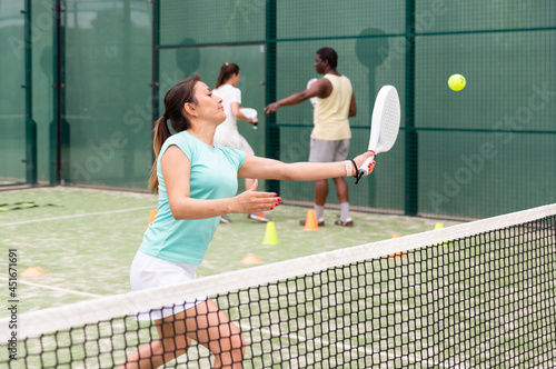 Happy woman learning to play padel game on tennis court outdoor. Other athletes are training in the background © JackF