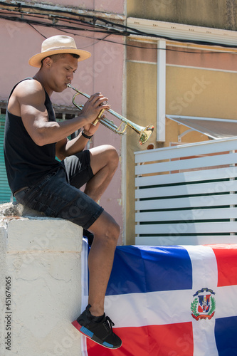 Latino man playing a trumpet sitting on a wall with the Dominican Republic flag: Selective focus.