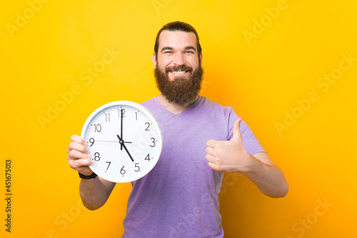 Happy bearded man is holding a round clock and showing thumb up.