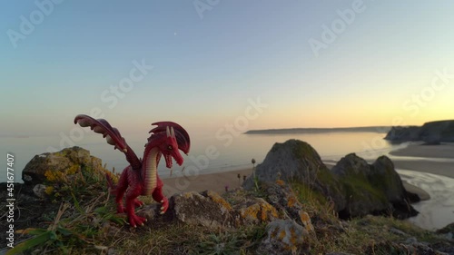 Red toy dragon overlooking Three Cliffs Bay at sunset, the Gower peninsula, Swansea, South Wales, United Kingdom. Symbol of Wales and Welsh landscape. Popular destination with beautiful beach.