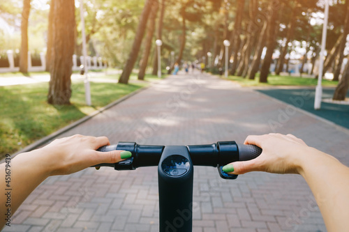 Young woman drives an electric scooter in a city park. Point of view on the steering wheel with female hands. photo