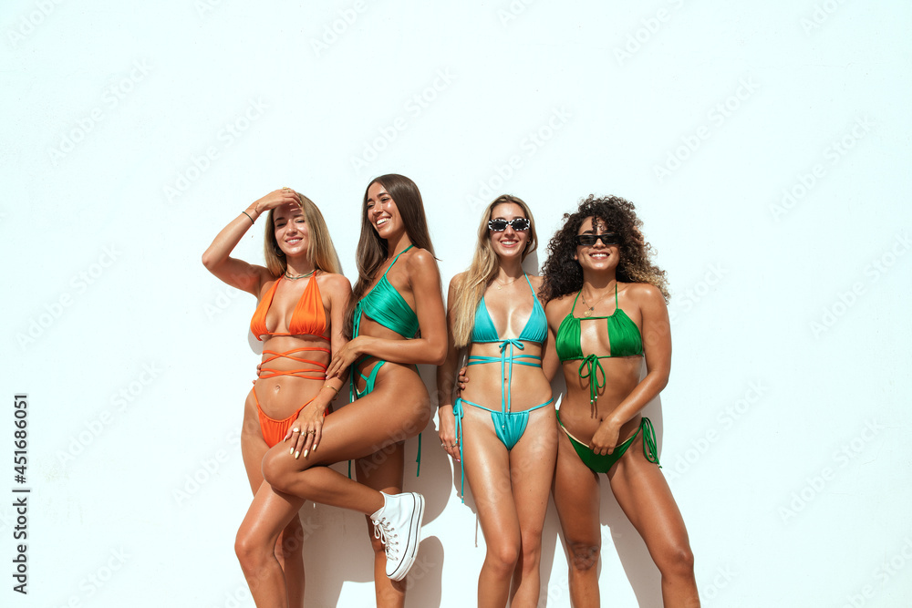 a group of multicultural girls in bikini of different colors laughing with a white wall behind