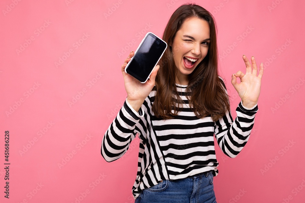 Photo of winking beautiful happy young woman wearing striped sweater isolated over background with copy space showing ok gesture looking at camera showing mobile phone screen Photo of winking beautiful happy young woman wearing striped sweater isolated over background with copy space showing ok gesture looking at camera showing mobile phone screen