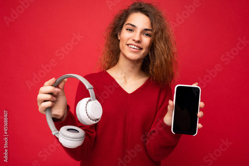 Charming funny smiling positive young brunette curly woman wearing dark red sweater isolated on red backdrop holding and suggesting white wireless headphones and mobile phone with empty screen for
