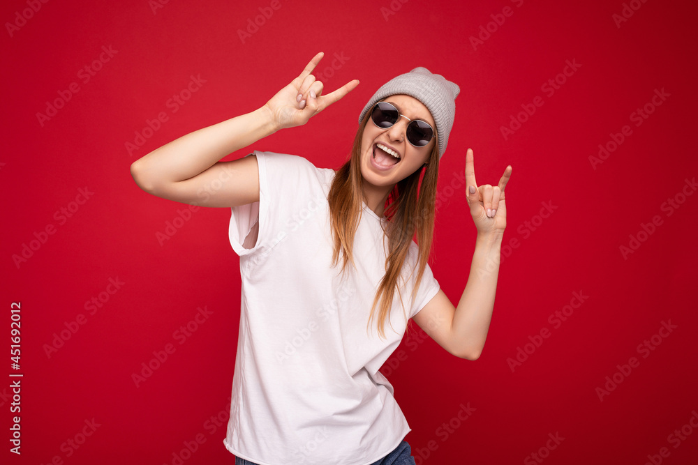 Photo shot of beautiful positive young dark blonde woman wearing casual clothes and stylish sunglasses isolated over colorful background wall looking at camera having fun and showing rock and roll Photo shot of beautiful positive young dark blonde woman wearing casual clothes and stylish sunglasses isolated over colorful background wall looking at camera having fun and showing rock and roll