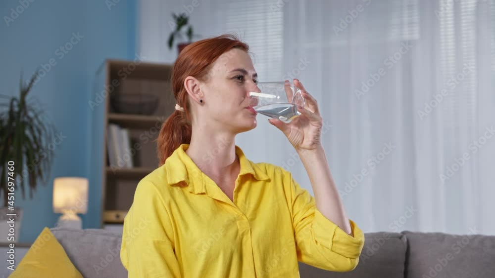 portrait of a young woman quenching her thirst and drinking cool clear water from a glass while sitting on the sofa in the room portrait of a young woman quenching her thirst and drinking cool clear water from a glass while sitting on the sofa in the room