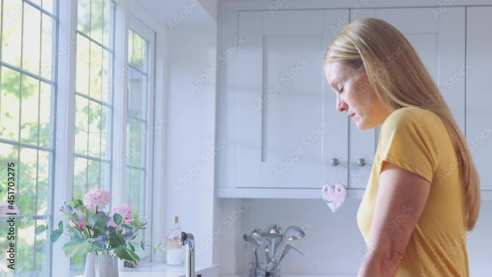 Depressed mature woman at home standing by kitchen window - shot in slow motion Depressed mature woman at home standing by kitchen window - shot in slow motion