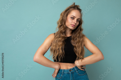 Young self-confident sexy beautiful winsome blonde curly woman with sincere emotions wearing stylish black top isolated over blue background with copy space © Ivan Traimak