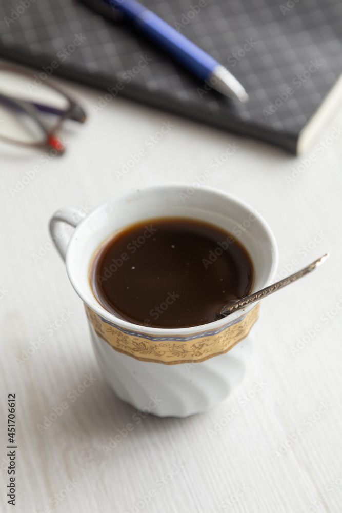 beautiful porcelain cup with coffee and a spoon inside, on a desk with glasses, a notebook and pen, drinks in the workplace, studio beautiful porcelain cup with coffee and a spoon inside, on a desk with glasses, a notebook and pen, drinks in the workplace, studio