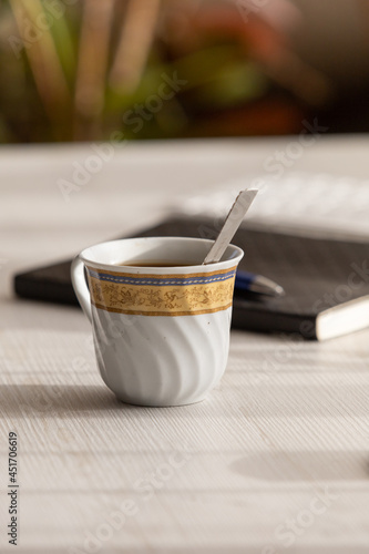 beautiful porcelain cup with coffee and a spoon, in the background is a notebook and pen, office objects in studio