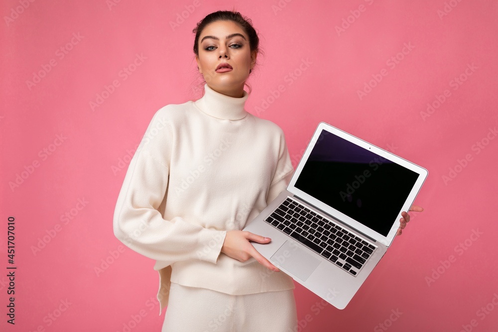 Photo of beautiful confudent arrogant brunet young woman with gathered dark hair wearing white sweater holding computer laptop and looking at camera isolated on pink wall background Photo of beautiful confudent arrogant brunet young woman with gathered dark hair wearing white sweater holding computer laptop and looking at camera isolated on pink wall background