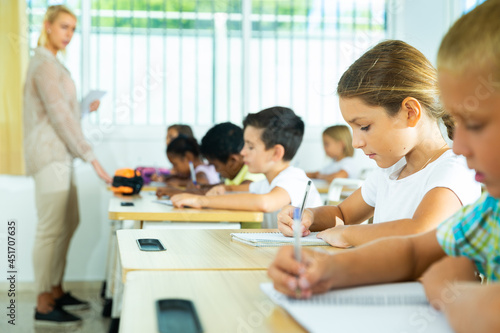 Diligent elementary school student tween girl studying with classmates  making notes of teacher lecture