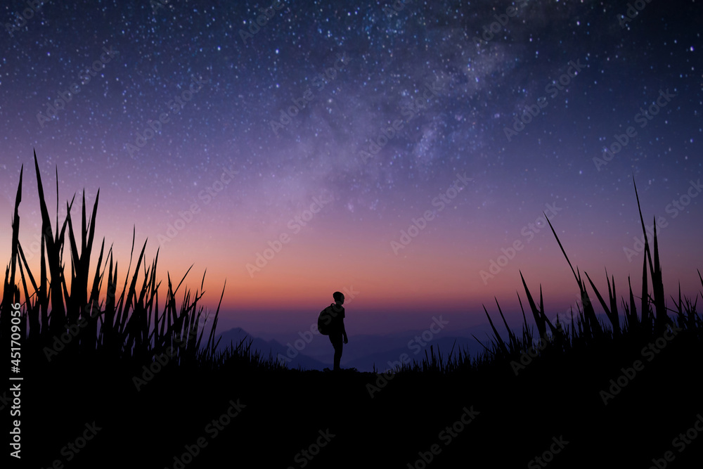 Silhouette of young traveler with backpack watched beautiful view of night sky, star, milky way alone on meadow over the mountain. He enjoyed traveling and was successful when he reached the summit.