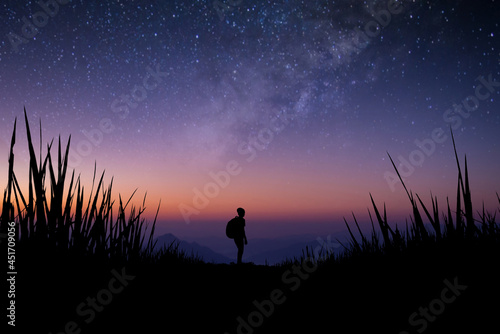 Silhouette of young traveler with backpack watched beautiful view of night sky, star, milky way alone on meadow over the mountain. He enjoyed traveling and was successful when he reached the summit.