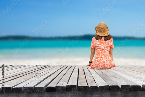 Summer beach vacation concept, Asia woman with hat sitting on wood floor on the beach look at the beauty of the white sandy beach, sea and sky. She felt happy and relaxed when she came to the beach. © Tarokmew