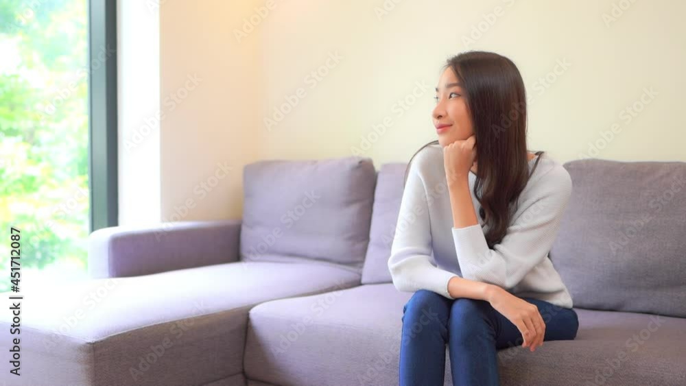 Serene Asian girl thinking while looking outside through the window, sitting near the window on the sofa during Thailand covid-19 lockdown Serene Asian girl thinking while looking outside through the window, sitting near the window on the sofa during Thailand covid-19 lockdown