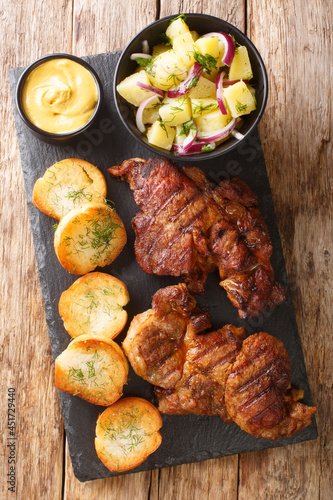 Authentic Schwenkbraten Marinated BBQ Pork neck steak with potatoes salad, mustard and toast close up in the slate board on the table. Vertical top view from above
