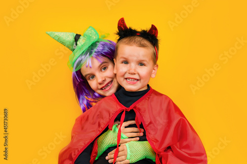 Cute girl kid in a witch costume with a green hat and a toddler boy with horns and a red raincoat in a little devil costume stands isolated on a yellow background.. Halloween, traditions concept.
