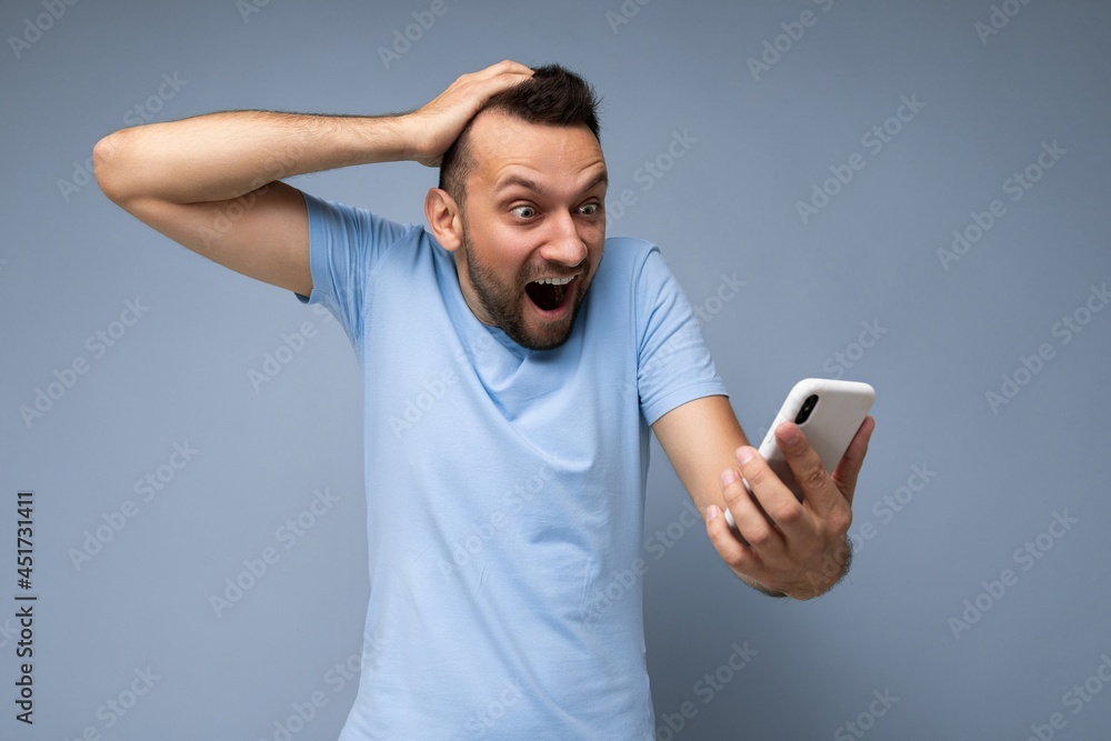 Furious shocked handsome young bearded brunet man wearing everyday blue t-shirt isolated over blue background holding and using mobile phone communication online on the internet looking at gadjet Furious shocked handsome young bearded brunet man wearing everyday blue t-shirt isolated over blue background holding and using mobile phone communication online on the internet looking at gadjet