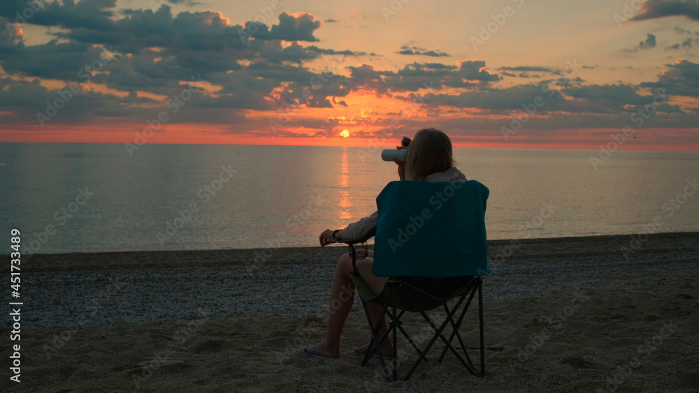 Back view of woman sitting on camping chair and drinking coffee. Relaxing and enjoying nature. Admiring sunrise on the sea.