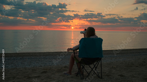 Back view of woman sitting on camping chair and drinking coffee. Relaxing and enjoying nature. Admiring sunrise on the sea. © art24pro
