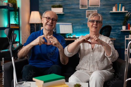 Caucasian old happy couple making heart sign with hands while looking at camera cheerful sitting in living room at home. Retired cheerful man and woman showing romantic love symbol