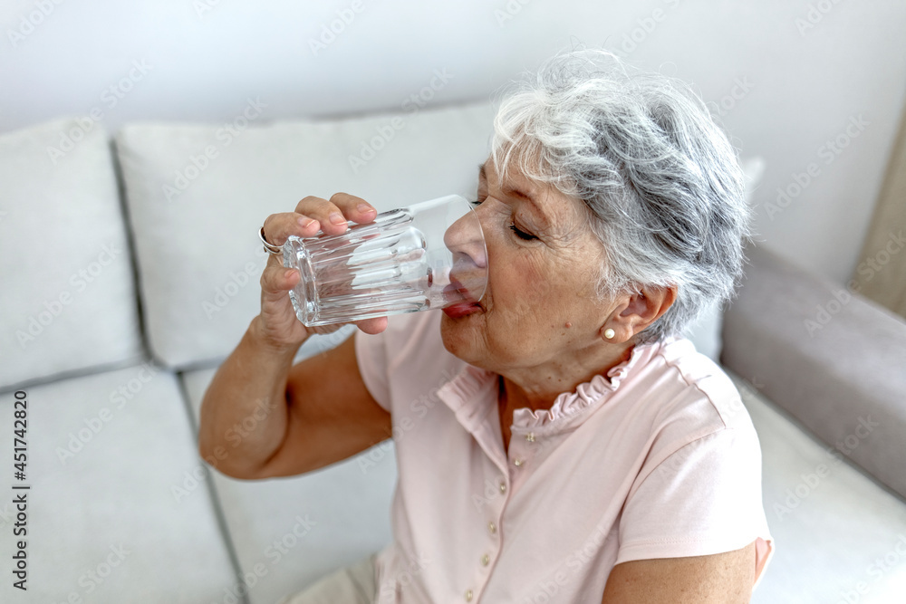 Beautiful old woman drinking water. Happy senior woman with glass of water at home. Thirsty woman drinking a refreshing healthy glass of cold fresh water. Age, health care and people concept.