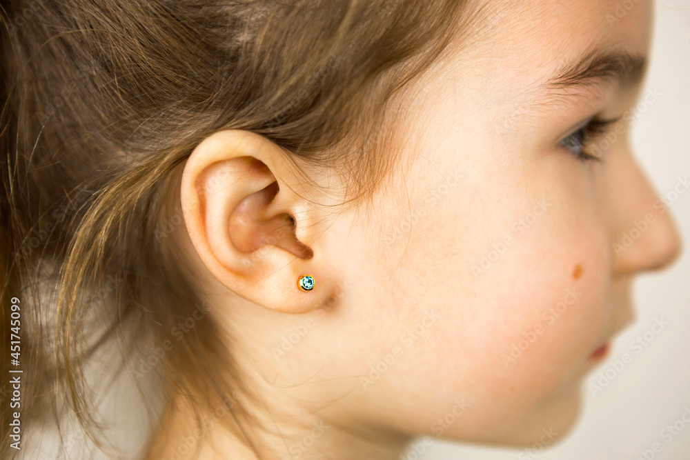 Ear piercing in a child - a girl shows an earring in her ear made of a medical alloy. White background, portrait of a girl with a mole on his cheek in profile.