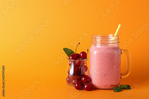 Glass jar of cherry smoothie and ingredients on orange background