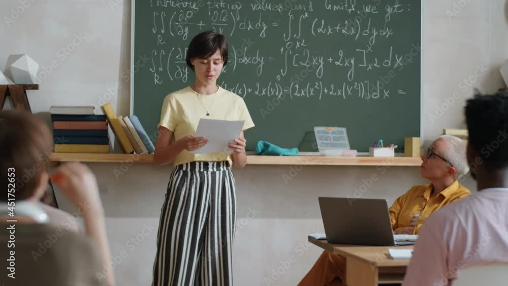 Young female student standing by chalkboard with formulas and reading aloud something on paper while reciting lesson in front of multiethnic classmates and teacher Young female student standing by chalkboard with formulas and reading aloud something on paper while reciting lesson in front of multiethnic classmates and teacher