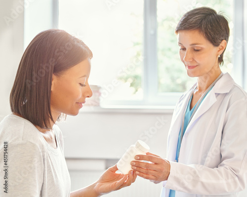 Smiling middle-aged woman doctor in white coat prescribing medicine to young african american female patient during medical healthcare appointment in clinic, physician giving vitamin bottle to client